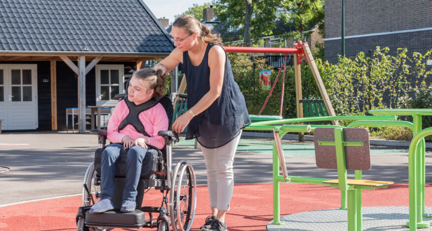 Eine Frau begleitet ein Kind mit Assistenzbedarf über den Spielplatz. Rechts im Bild ist ein Rollstuhlkarusell zu sehen.